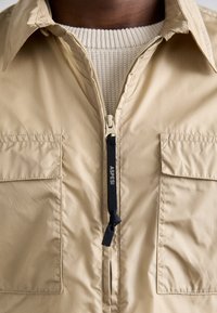 Veste beige avec fermeture éclair frontale étiquetée "Aspesi" sur un pull tricoté crème, montrant le col et la partie supérieure de la poitrine.