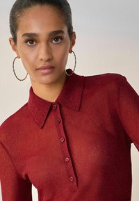 Rotes, geripptes Langarmshirt mit Kragen, ausgestattet mit einer Knopfleiste und subtilen Glitzereffekten. Große Creolenohrringe ergänzen das Design.