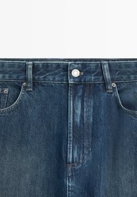 Jeans in denim blu scuro con davanti con bottone, chiusura lampo, passanti per cintura e tasche frontali.