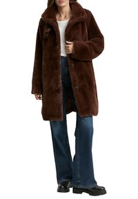 Brauner Kunstpelzmantel mit großem Kragen, Knopfverschluss und weiten Ärmeln, kombiniert mit ausgestellten Jeans und schwarzen Plateau-Stiefeln.