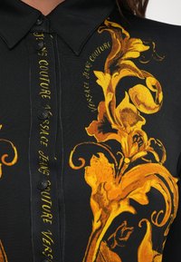 Juodas marškinėlis su sagomis, puošniais geltonais ir oranžiniais baroko gėlių raštais, o prie apykaklės ir dizaino matyti "Versace Jeans Couture" tekstą.