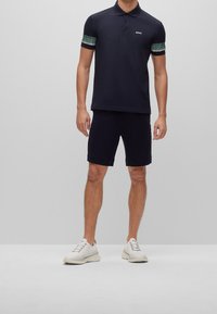 Polo marine avec des accents verts à motifs sur les manches, associé à un short noir et des baskets beiges. Présente un petit logo sur la poitrine.