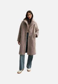 Lichtbruin faux shearling jas met een brede kraag, twee knopen en zijzakken, gedragen over een crèmekleurige trui en blauwe spijkerbroek.