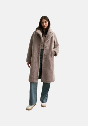 Light brown faux shearling coat with a wide collar, two buttons, and side pockets, worn over a cream sweater and blue jeans.