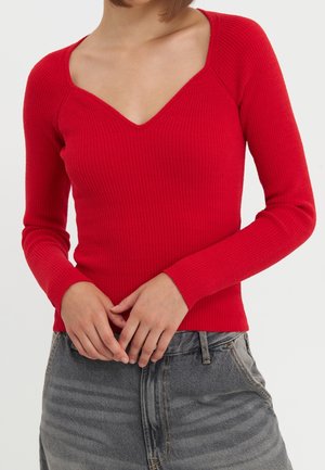 Femme portant un haut rouge moulant côtelé à manches longues avec un col en V profond et un jean gris taille haute, mains jointes devant.