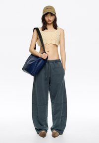 Háčkovaný crop top v béžovej farbe, široké šedé tepláky s detailmi šitia, námornícka kožená kabelka cez rameno a béžová čiapka s logom.