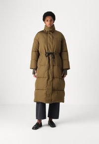 Lang olijfgroen pufferjack met een getailleerde taille, hoge kraag en een ontwerp met verlaagde schouders. Heeft een horizontaal gestikt patroon en een trekkoord.