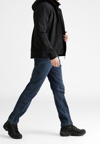 Veste noire zippée avec capuche, jeans en denim bleu et bottes de randonnée noires. La veste présente des poches zippées et une texture lisse.
