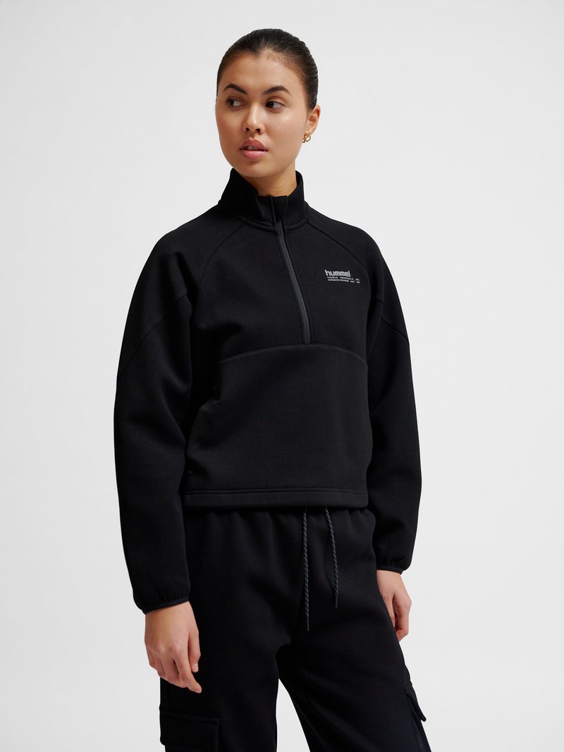 Vrouw met zwarte trui met kwartrits en bijpassende zwarte broek, kijkt naar links tegen een effen lichte achtergrond.