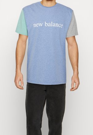 Mężczyzna w kolorowej koszulce New Balance z niebieskim korpusem, miętowozielonym lewym rękawem, szarym prawym rękawem oraz czarnych spodniach.