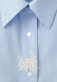 Svetlomodrá košeľa s golierom, bielymi gombíkmi a malou textúrovanou bielou kvetinovou výšivkou pri strednom zapínacom páse.
