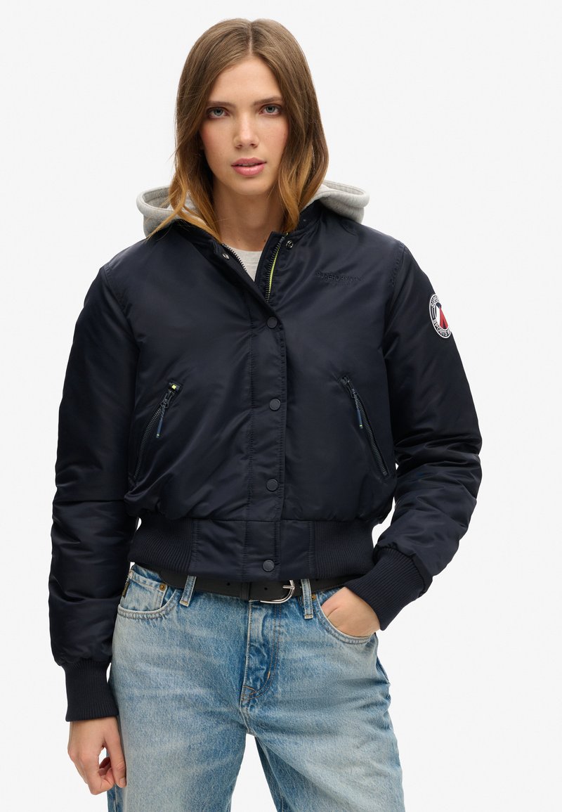 Marine bomberjakke med grå hætte, frontlomme med lynlås, ribbede manchetter og kant, samt logo-patch på armen. Struktureret materiale, cropped fit.
