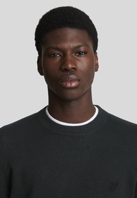 Suéter de punto verde oscuro con un cuello redondo y un interior de color blanco, con un pequeño logo oscuro en el pecho. Textura suave.