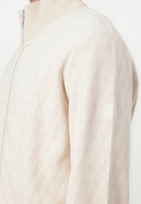 Veste zippée beige avec un motif de feuilles géométriques, col côtelé et un petit logo "OPEN" en blanc sur la manche.