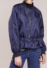 Chaqueta bomber de satén azul marino con cuello acanalado, cintura elástica, cierre de cremallera y detalle de volantes en el dobladillo. Incluye bolsillos laterales.