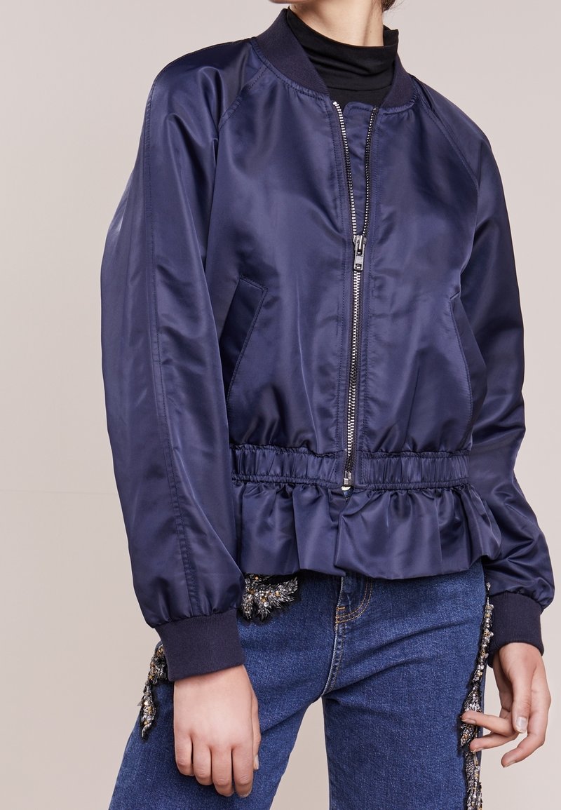 Chaqueta bomber de satén azul marino con cuello acanalado, cintura elástica, cierre de cremallera y detalle de volantes en el dobladillo. Incluye bolsillos laterales.