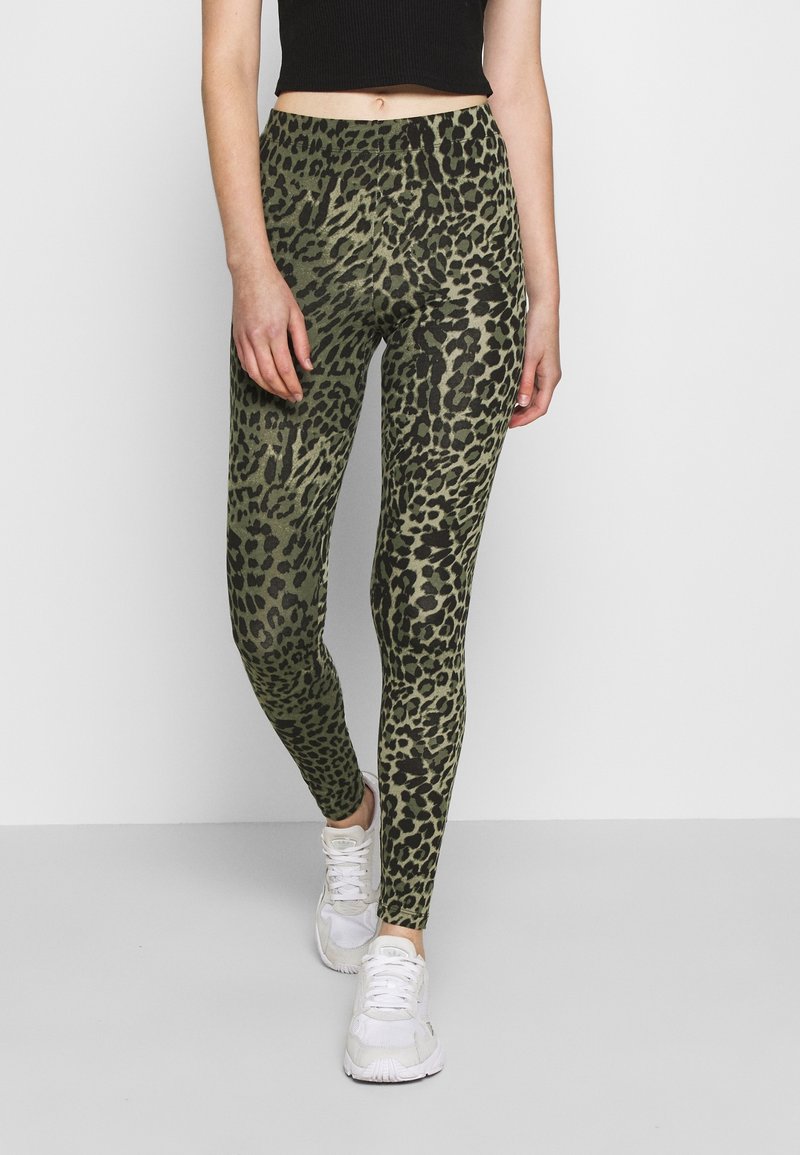 Mujer vistiendo leggings de estampado de leopardo verde y negro de cintura alta, combinados con zapatillas blancas y un top corto negro .