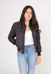 Czarna pikowana kurtka bomber z złotym zamkiem błyskawicznym, żebrowymi mankietami i pomarańczową metką przy dolnym brzegu, zestawiona z białą bluzką i niebieskimi dżinsami.