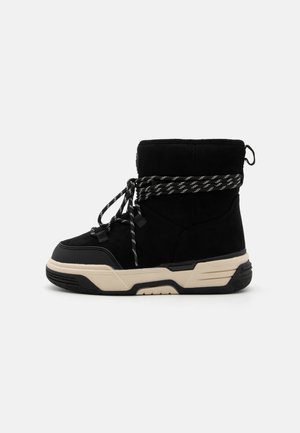 Bottines en suede noir avec un embout texturé, une semelle intermédiaire beige contrastante, et des lacets en nylon noir et blanc tressés. Avec une languette à l'arrière.