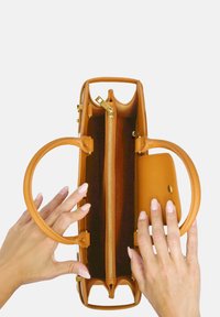 Tan leren handtas met ronde handvaten, ritssluiting en een kleinere voorkantzak. Het interieur heeft een gestructureerde voering en gouden hardware.