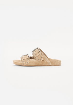 Tan suede slide sandals with two wide straps, featuring metal buckles and decorative eyelet holes. Flat sole with a textured finish.