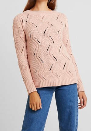 Light pink knitted sweater with wave patterns, textured stitching, and ribbed cuffs. Worn with blue denim jeans.