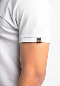 Biela polo košeľa s textúrovaným povrchom a krátkymi rukávmi. Na leme rukáva sa nachádza čierna visačka so žltým logom.
