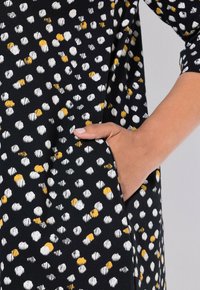 Tessuto nero con pattern di punti bianchi e gialli sparsi. Il capo presenta una tasca laterale e una superficie liscia e testurizzata.