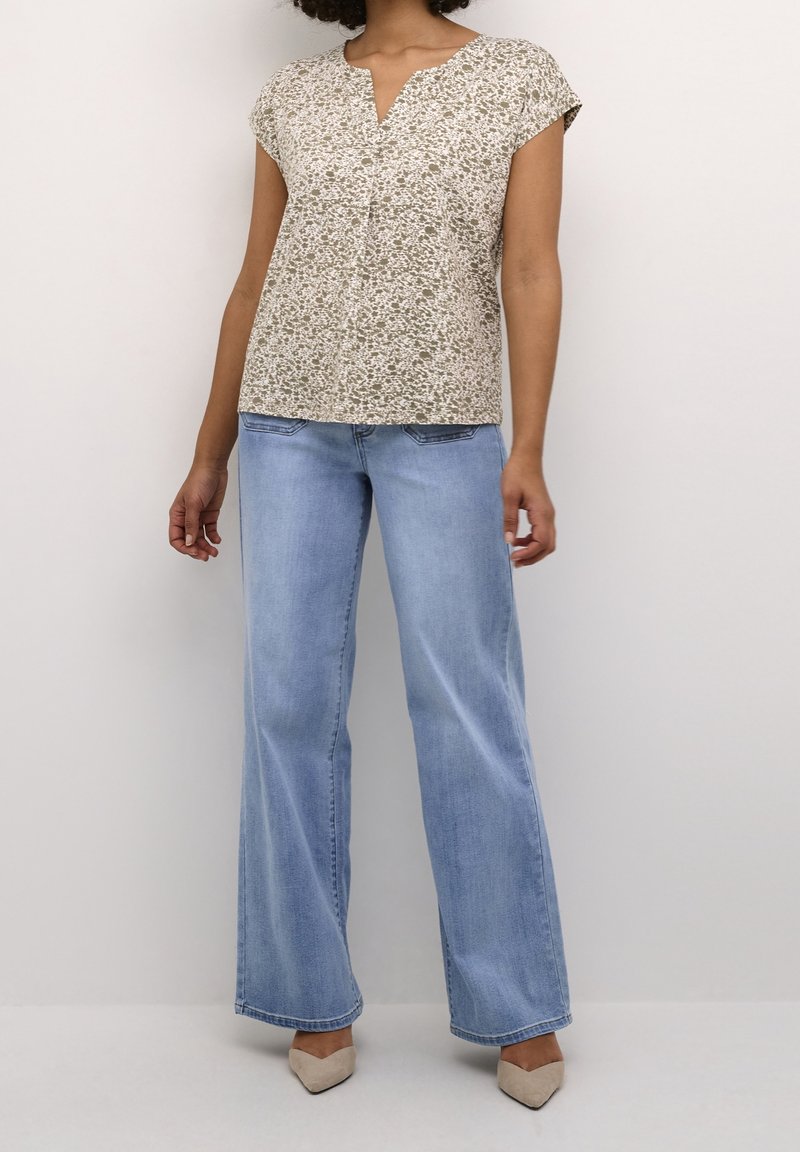 Femme portant un chemisier beige à manches courtes à motifs, un jean large bleu clair et des chaussures beige à bout pointu, debout devant un fond uni.