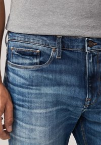 Primer plano de una persona que lleva jeans de mezclilla azules desgastados y una camisa gris clara, con una mano relajada a su lado.