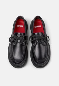 Black leather shoes with a rounded shape. Features a thick black sole, grey laces, and red insoles printed with "CAMPER." Smooth texture.