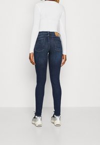 Mörkblå slim fit-jeans med hög midja, med två bakfickor och en läderetikett på midjebandet, tillsammans med en vit långärmad topp.