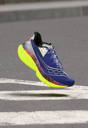 Jooksujalats sinise võrgust ülaosaga, mustade ja oranžide detailide, lilla paelte ning paksu erksakollase tallaga, mis hõljub asfaltteekonna kohal.