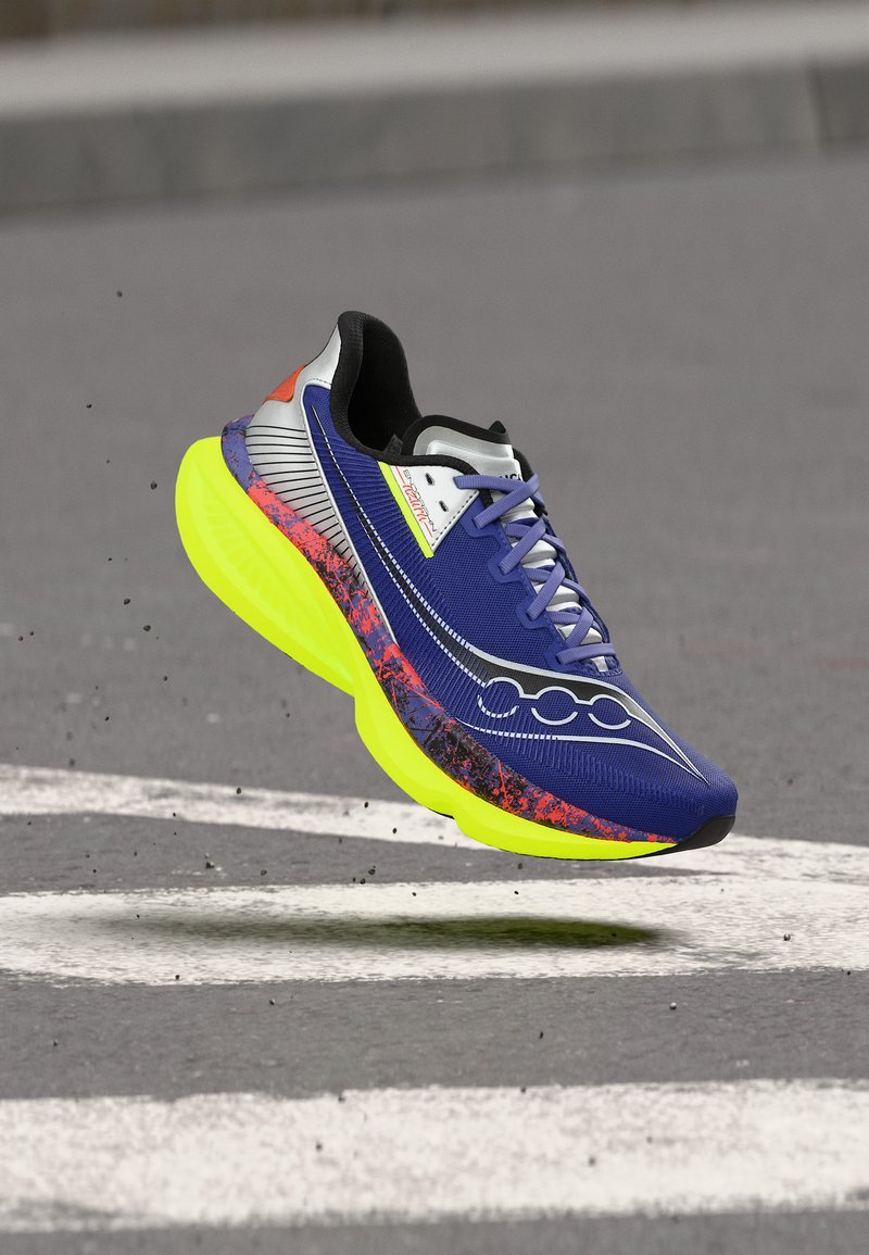 Αθλητικό παπούτσι με μπλε διχτυωτό επάνω μέρος, μαύρες και πορτοκαλί λεπτομέρειες, μωβ κορδόνια και παχιά φωτεινή κίτρινη σόλα που αιωρείται πάνω από ασφαλτοστρωμένο δρόμο.