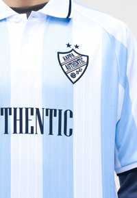 Αθλητική μπλούζα με κάθετες ρίγες σε ανοιχτό μπλε και λευκό χρώμα, με μαύρο λογότυπο "Kappa Authentic 1967" και κείμενο "AUTHENTIC" στο μπροστινό μέρος.