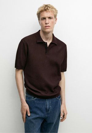 Junger Mann mit kurzen blonden Haaren, der ein dunkelbraunes kurzärmeliges Polohemd und blaue Jeans vor einem schlichten weißen Hintergrund trägt.