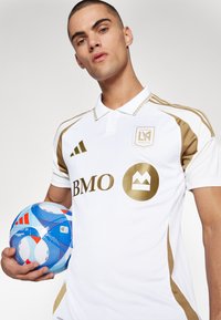 Camiseta de fútbol blanca con acentos dorados y logo, que presenta un cuello y paneles laterales. El jugador sostiene un balón de fútbol con un patrón azul y rojo.