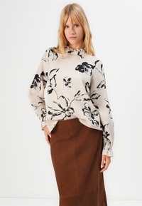 Donna bionda che indossa una blusa beige con grandi stampe floreali nere e colletto alto, abbinata a una gonna marrone, in piedi su uno sfondo bianco.