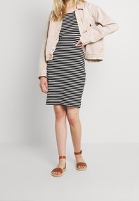 Veste en denim rose pâle portée sur une robe rayée noir et blanc, associée à des sandales marron. Les détails incluent des boutons sur la veste et des lanières autour de la cheville.