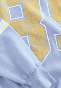 Felpa azzurra chiara con un grande numero giallo "98" contornato di bianco, texture morbida, polsini a coste e strisce a contrasto sulle spalle.