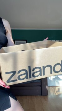 Kartonowe pudełko z napisem "zalando" wypisanym dużymi czarnymi literami. Pudełko jest otwarte, ukazując ciemne przedmioty w środku. W tle znajduje się sofa i ściana.
