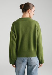 Zielony dzianinowy sweter z szerokim, prążkowanym dołem i luźnym krojem. Prezentowany na osobie zwróconej tyłem, w połączeniu z jasnoniebieskimi dżinsami.