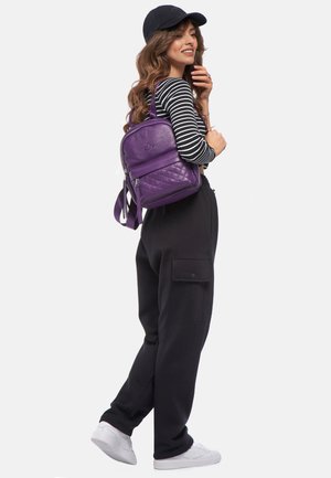 Lila, gesteppte Rucksack mit doppeltem Reißverschluss, an einer Schulter hängend und mit einer glatten Textur gestaltet. Gestreiftes Langarmshirt und schwarze Hose.