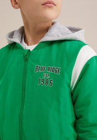 Zeleni bomber jakna sivi hoody oblogu. Ima bijele detalje i izvezeni natpis: "BLUE RIDGE EST. 1986" je na prednjoj strani. Zatvaranje na munju.