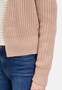 Gebreide cardigan in zacht roze, met een grof textuur, geribbelde mouwen en een klein metalen accent. Gecombineerd met blauwe denim.