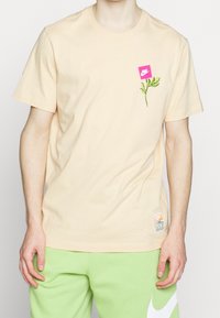 Jasno brzoskwiniowy bawełniany t-shirt z okrągłym dekoltem, z różowym kwadratowym logo i zieloną grafiką liścia na lewej piersi.