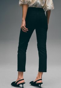 Černé džíny s vysokým pasem, úzkým střihem a rovnými nohavicemi, se zadními kapsami. Nosené s černými slingback podpatky a bílou blůzou se vzorem.