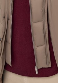 Beżowa kurtka puchowa z zamykanymi kieszeniami, noszona na bordowym swetrze z dzianiny. Teksturowana tkanina o gładkim wykończeniu, widoczne części jasnych spodni.