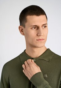 Grünes Langarm-Polohemd mit Kragen, ausgestattet mit einem gestickten Logo, strukturiertem Stoff und zwei Knöpfen am Hals.