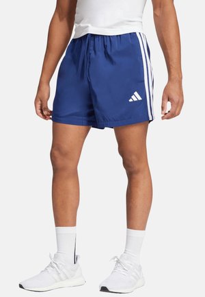 Männlich, der blaue Adidas-Shorts mit weißen Streifen, weiße Socken, weiße Sneakers und ein weißes eng anliegendes ärmelloses Shirt trägt.